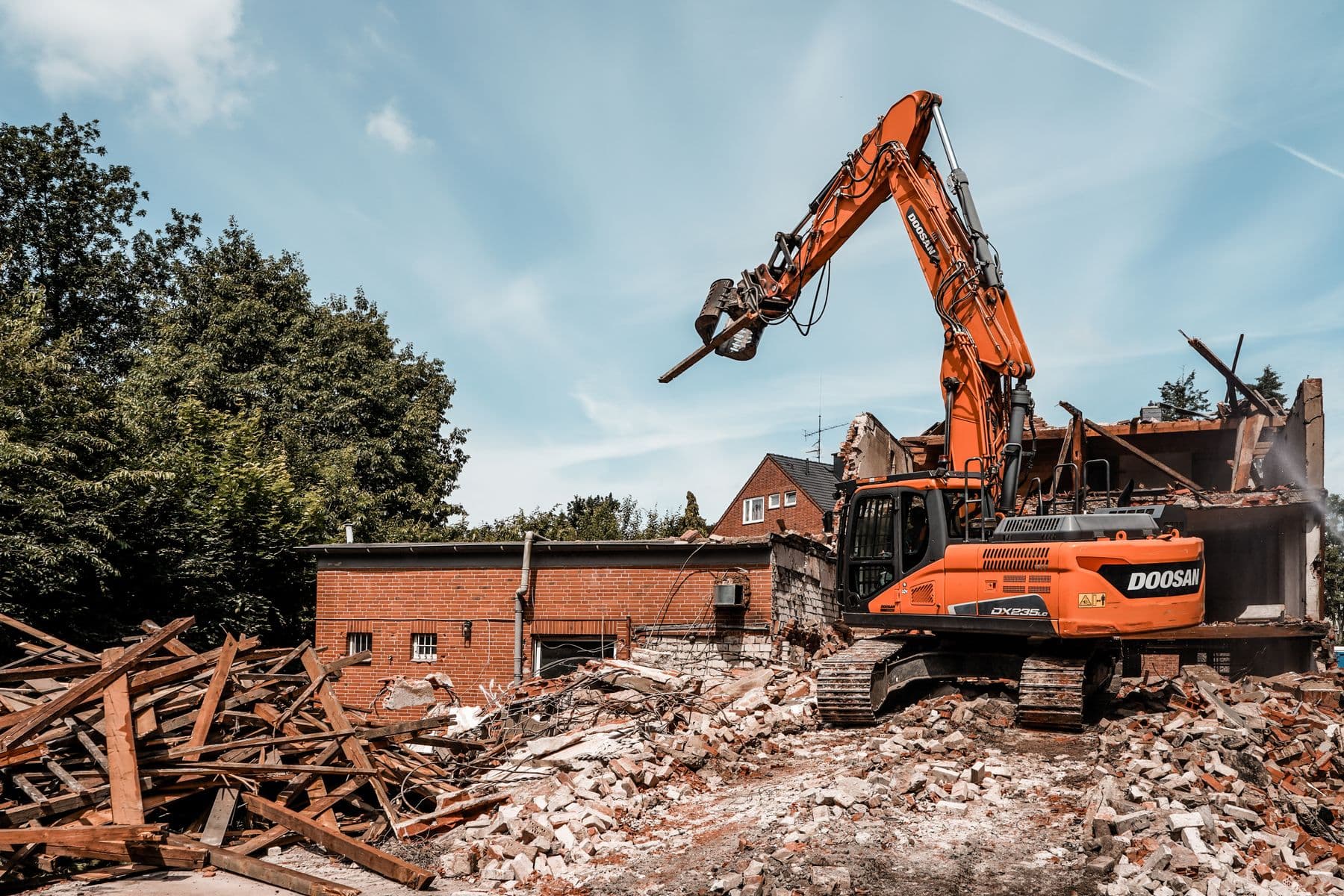 An orange excavator pulling down a brick building, with timber and rubble in front of it.